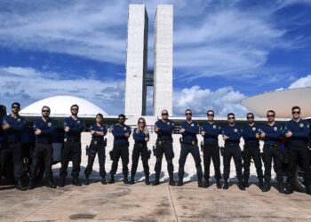 Grupo de agentes federais em uniforme azul posando com braços cruzados diante do Congresso Nacional do Brasil