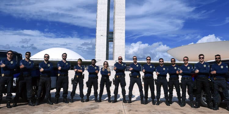 Grupo de agentes federais em uniforme azul posando com braços cruzados diante do Congresso Nacional do Brasil