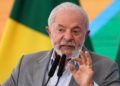 Presidente atual do Governo, Lula Inácio, discursando com veemência diante de microfones, bandeira brasileira ao fundo.