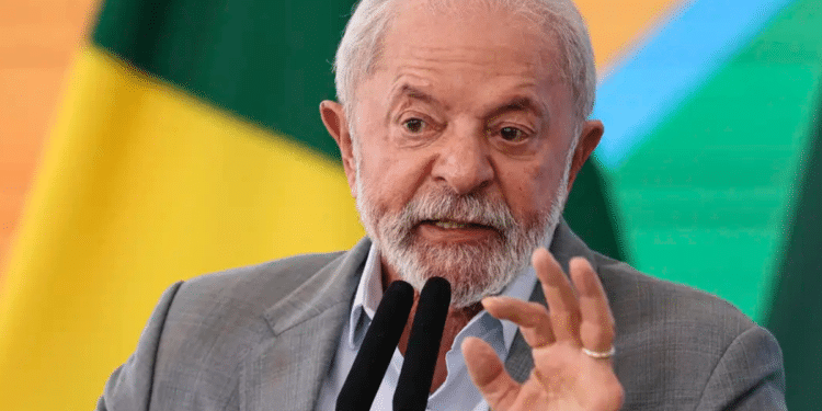 Presidente atual do Governo, Lula Inácio, discursando com veemência diante de microfones, bandeira brasileira ao fundo.