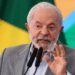 Presidente atual do Governo, Lula Inácio, discursando com veemência diante de microfones, bandeira brasileira ao fundo.