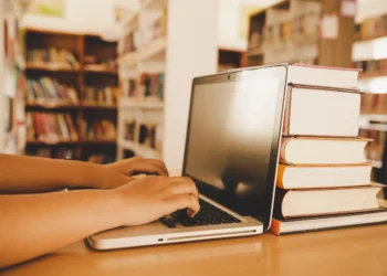 Pessoa utilizando notebook em biblioteca para acompanhar processos seletivos na área da Educação com inscrições abertas.