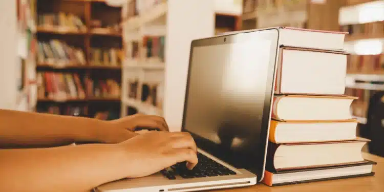 Pessoa utilizando notebook em biblioteca para acompanhar processos seletivos na área da Educação com inscrições abertas.