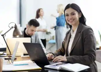 Mulher sorrindo enquanto trabalha em um laptop em um ambiente de escritório moderno.