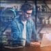 Homem jovem de óculos e camisa jeans trabalhando em um notebook, cercado por interfaces digitais holográficas azuis que exibem gráficos, dados e ícones de tecnologia.
