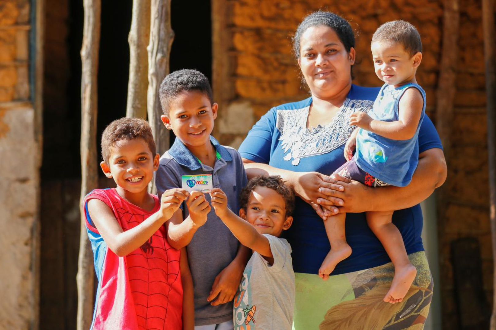 Família beneficiária do programa Mães de Pernambuco. Mulher com quatro filhos segurando o cartão do programa Mães de Pernambuco, com fundo de casa simples.