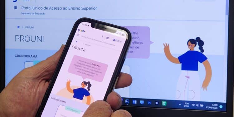 Close de mão usando smartphone com tela do Prouni, notebook com portal Prouni ao fundo.