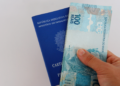 Mão segurando uma nota de 100 reais em frente a uma carteira de trabalho azul do Brasil