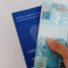 Mão segurando uma nota de 100 reais em frente a uma carteira de trabalho azul do Brasil
