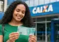 Mulher segura uma nota de R$ 100 em frente a uma agência da Caixa, representando o ressarcimento do PIS/Pasep.