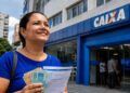 Mulher segura dinheiro e documento em frente a uma agência da Caixa, em referência ao saque de mais de R$ 6 mil do FGTS.