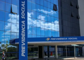 Fachada do prédio do INSS com o nome 'Previdência Social' visível e um céu refletido nas janelas.
