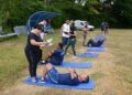 Pessoas realizando exercícios abdominais em colchonetes, sendo avaliadas por instrutores ao ar livre em 2026