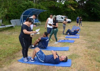 Pessoas realizando exercícios abdominais em colchonetes, sendo avaliadas por instrutores ao ar livre em 2026