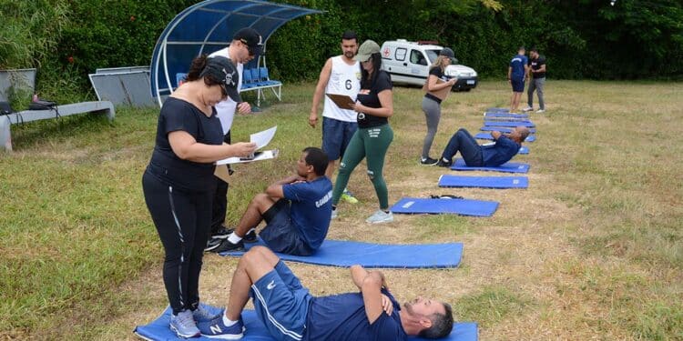 Pessoas realizando exercícios abdominais em colchonetes, sendo avaliadas por instrutores ao ar livre em 2026