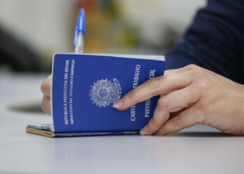 Mão segurando uma carteira de trabalho azul do Brasil sobre mesa branca com caneta azul ao fundo
