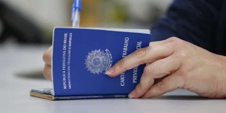 Mão segurando uma carteira de trabalho azul do Brasil sobre mesa branca com caneta azul ao fundo