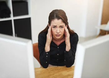 Mulher jovem e estressada, com expressão de frustração, sentada em sua mesa de trabalho, refletindo o estresse e arrependimento profissional após a graduação.