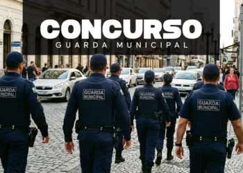 Vista traseira de cinco agentes da Guarda Municipal caminhando em formação por uma rua de paralelepípedos no centro da cidade. Eles vestem uniformes azuis escuros com a inscrição "GUARDA MUNICIPAL" em cinza nas costas e portam equipamentos de segurança na cintura.
