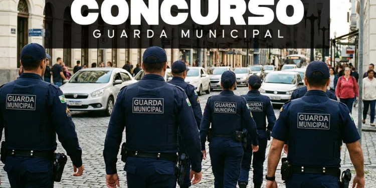 Vista traseira de cinco agentes da Guarda Municipal caminhando em formação por uma rua de paralelepípedos no centro da cidade. Eles vestem uniformes azuis escuros com a inscrição "GUARDA MUNICIPAL" em cinza nas costas e portam equipamentos de segurança na cintura.