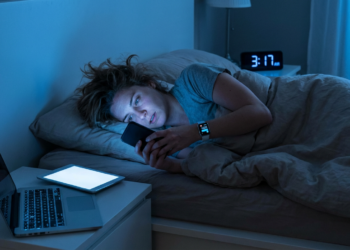 Mulher deitada na cama olhando o celular, cercada por notebook e tablet com tela acesa às 3h17.