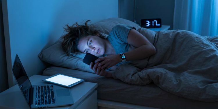 Mulher deitada na cama olhando o celular, cercada por notebook e tablet com tela acesa às 3h17.