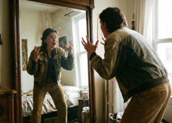 Jovem faz gestos dramáticos com as mãos em frente a espelho grande em quarto iluminado.