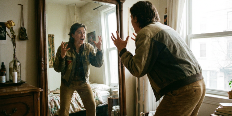 Jovem faz gestos dramáticos com as mãos em frente a espelho grande em quarto iluminado.