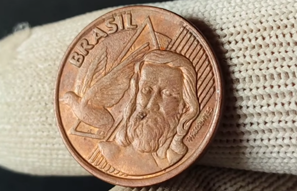 erro cunhagem moeda 5 centavos tiradentes duplicado Close do anverso de uma moeda de 5 centavos de bronze, segurada por dedos com luvas brancas. A imagem mostra a efígie de Tiradentes, um triângulo e uma pomba à esquerda, com a palavra "BRASIL" no topo. No canto inferior direito, o nome "TIRADENTES" aparece gravado de forma discreta.
