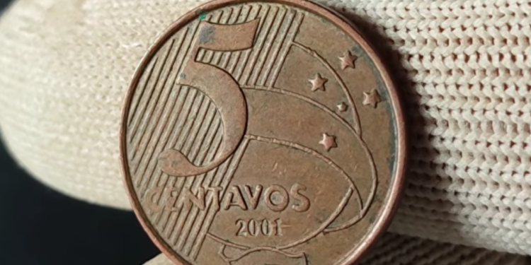 Close de uma moeda de 5 centavos do ano 2001 sendo segurada por dedos que usam luvas de tecido branco. A moeda de cobre mostra o número 5, a palavra "CENTAVOS" e o ano 2001 em destaque.