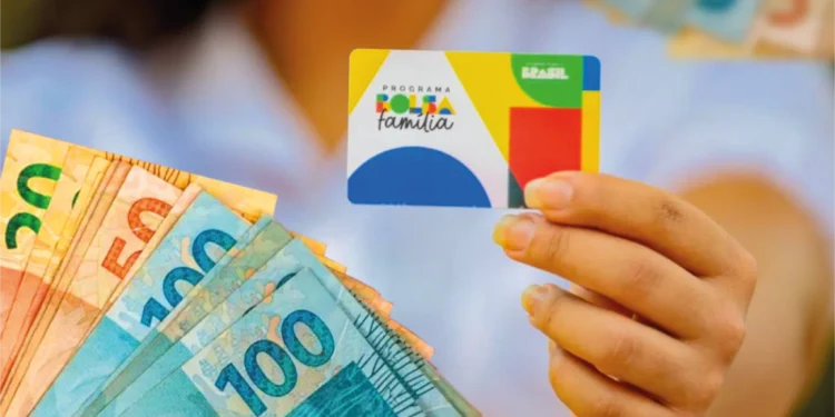Cartão do Programa Bolsa Família sendo segurado por uma mão, com notas de 20, 50 e 100 reais ao fundo, representando os pagamentos de março.