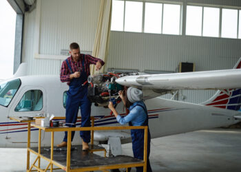 Dois profissionais trabalhando na manutenção de aeronave, um em cima de uma plataforma e outro na frente do motor da aeronave dentro de um hangar.
