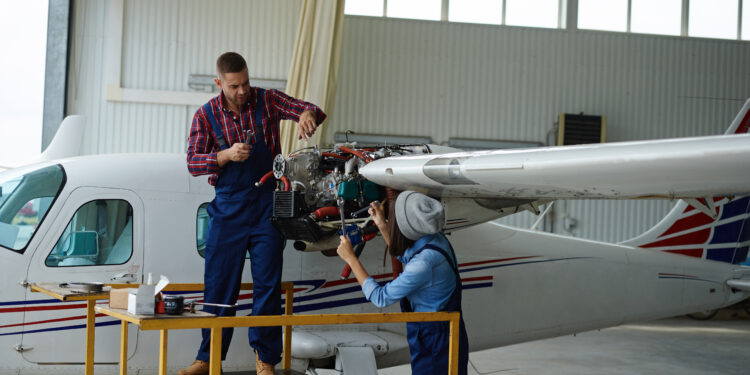 Dois profissionais trabalhando na manutenção de aeronave, um em cima de uma plataforma e outro na frente do motor da aeronave dentro de um hangar.