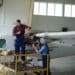 Dois profissionais trabalhando na manutenção de aeronave, um em cima de uma plataforma e outro na frente do motor da aeronave dentro de um hangar.