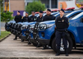 Guardas municipais perfilados ao lado de viaturas azuis em estacionamento urbano