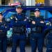 Quatro guardas municipais uniformizados posam com braços cruzados diante de viatura azul