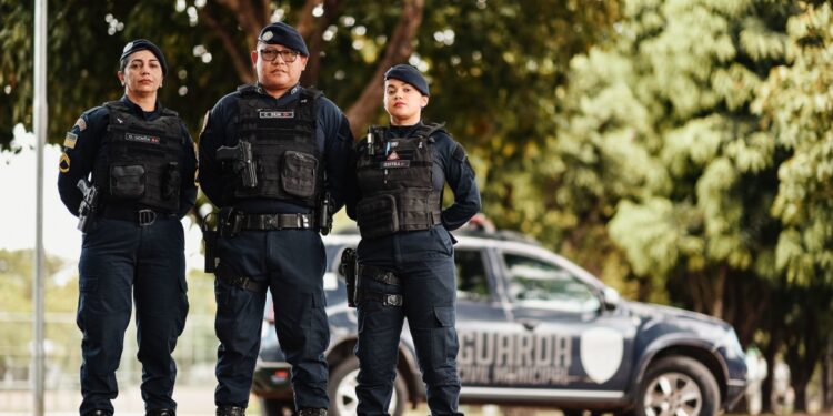 Três guardas civis municipais uniformizados em frente a viatura estacionada