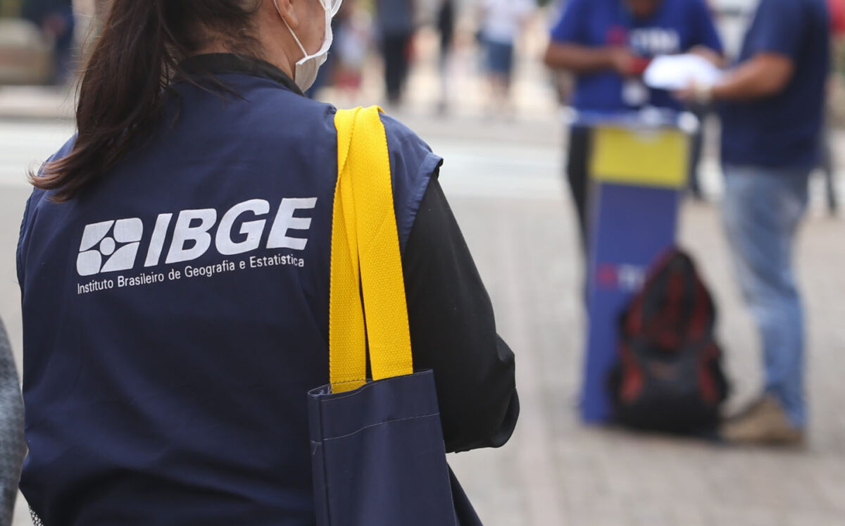 Concurso IBGE Mulher de colete do IBGE usa máscara enquanto segura bolsa amarela em rua movimentada