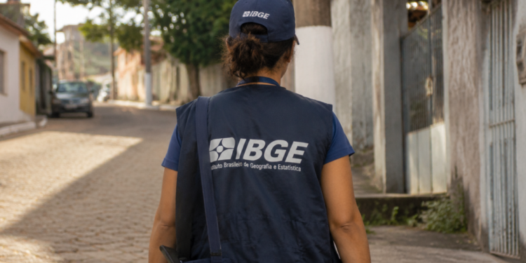 Recenseador do IBGE caminhando em rua residencial com uniforme durante coleta de dados para o Censo Agropecuário, Florestal e Aquícola.