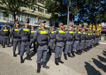 Grupo de policiais militares em uniforme cinza alinhados em praça durante solenidade