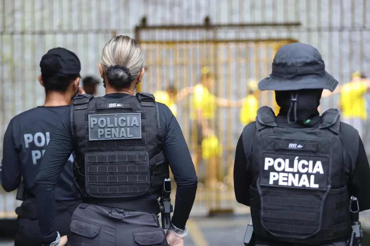 Concurso Polícia Penal Policiais penais de costas observam detentos com uniformes amarelos atrás de grades
