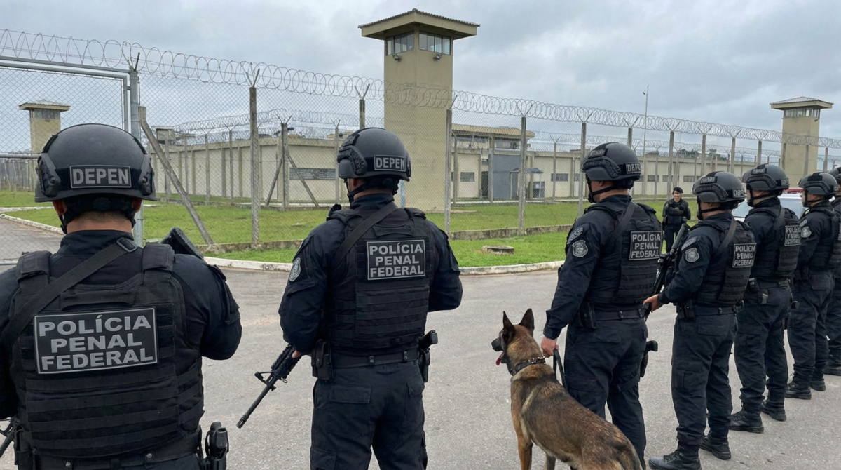 Concurso Polícia Penal Federal Grupo de policiais penais federais armados com cão de guarda em frente a presídio cercado por grades e arame farpado
