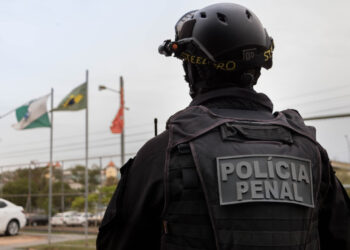 Agente da Polícia Penal observa área externa de presídio com bandeiras ao fundo