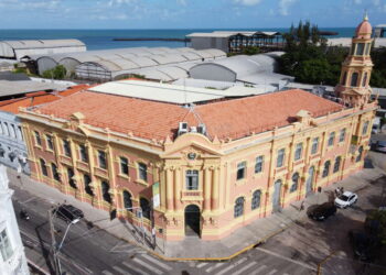 Edifício antigo com fachada amarela e rosa em esquina, próximo ao mar azul e céu parcialmente nublado