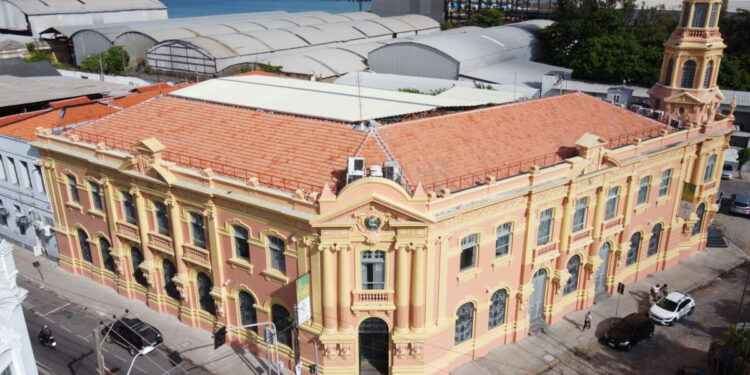 Edifício antigo com fachada amarela e rosa em esquina, próximo ao mar azul e céu parcialmente nublado