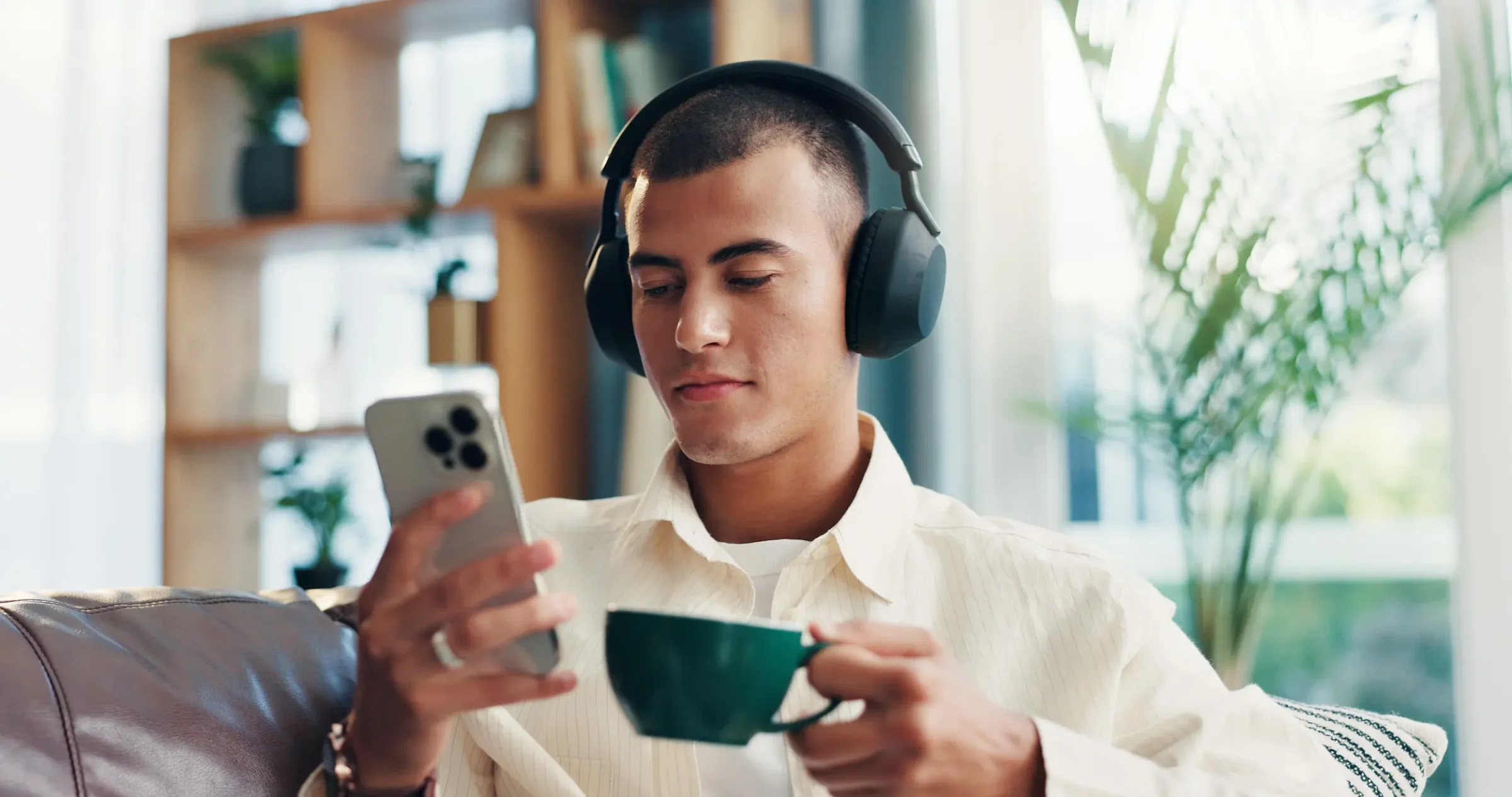 concursos 2026 Jovem estudando de forma ativa com fones de ouvido, assistindo a conteúdo no celular enquanto toma café. Representa o uso de mídias audiovisuais para otimizar o estudo durante o dia.