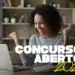 Mulher sorri e ergue os punhos ao lado de notebook, com livros ao fundo