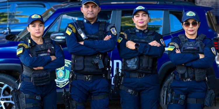 Quatro guardas municipais uniformizados posam de braços cruzados diante de viatura azul