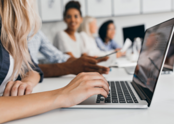 Mão digitando em notebook enquanto outros alunos acompanham aula presencial