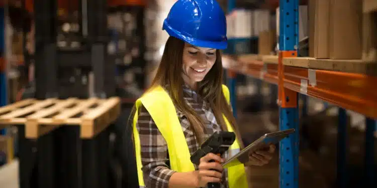 Mulher com capacete e colete de segurança analisando dados em um ambiente industrial, representando cursos técnicos em logística.